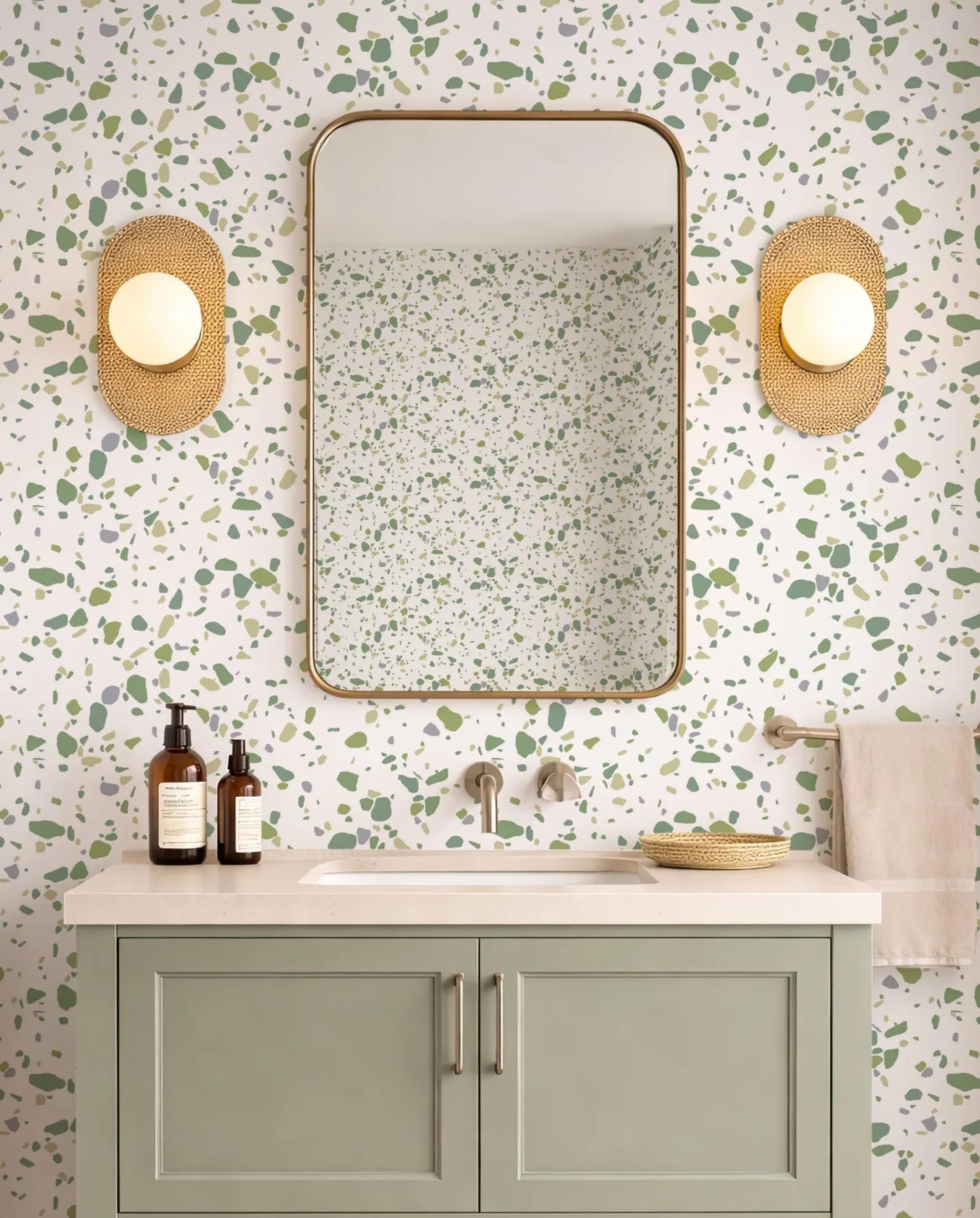 a bathroom with a white and green speckled wall, a white sink, a mirror, and two round wall sconces. The bathroom has a green cabinet below the sink and a beige towel hanging on the right side.