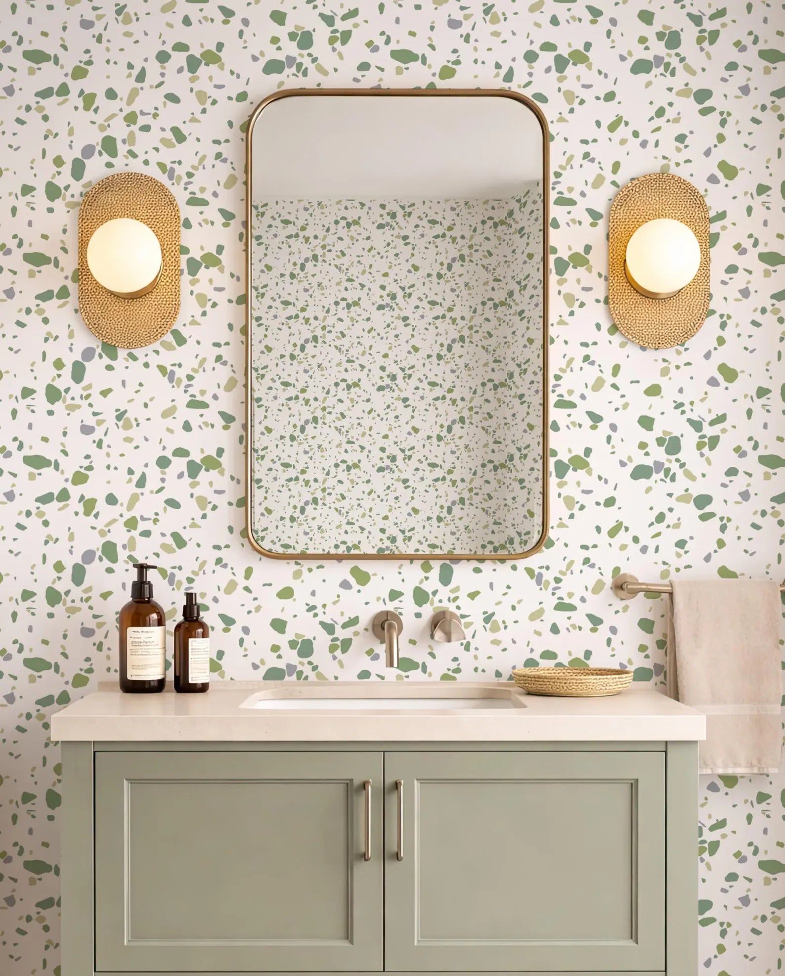 a bathroom with a white and green speckled wall, a white sink, a mirror, and two round wall sconces. The bathroom has a green cabinet below the sink and a beige towel hanging on the right side.