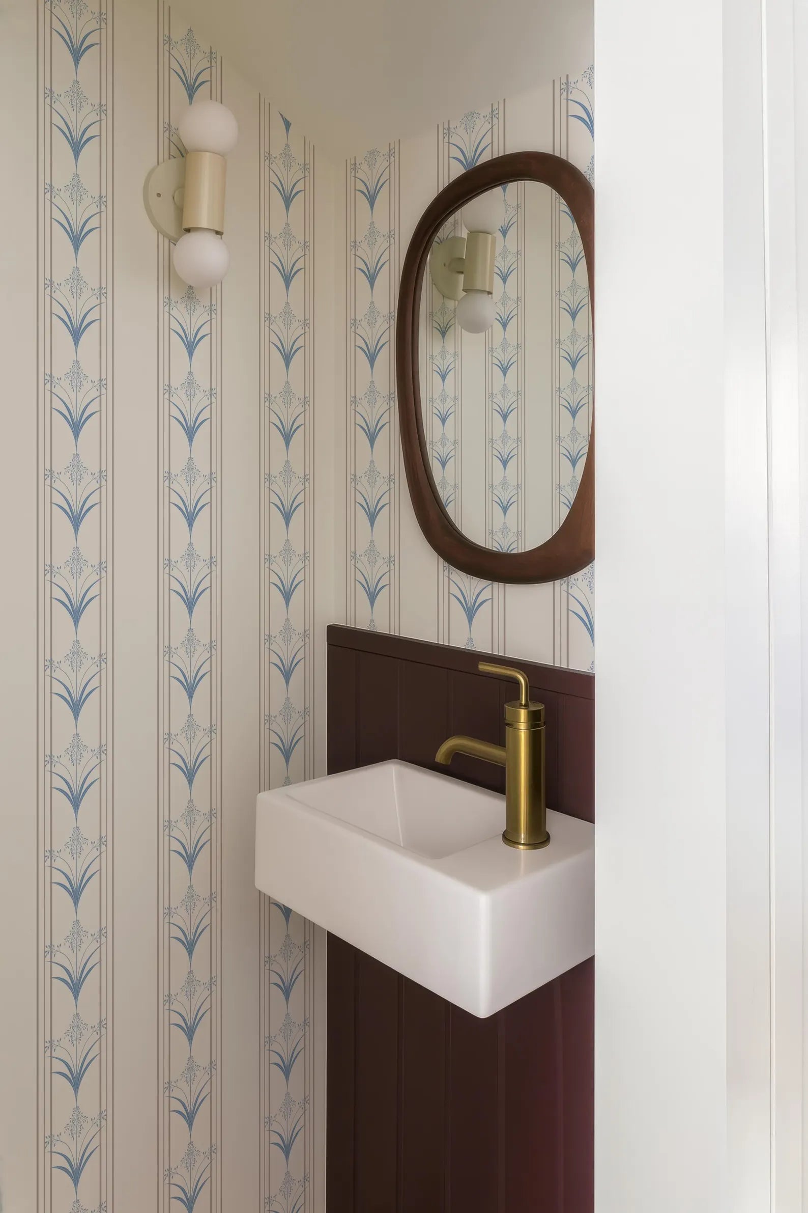 a bathroom with a white sink and a gold faucet, set against a wall with blue and white patterned wallpaper. There is a mirror above the sink, and a light fixture is mounted on the wall.