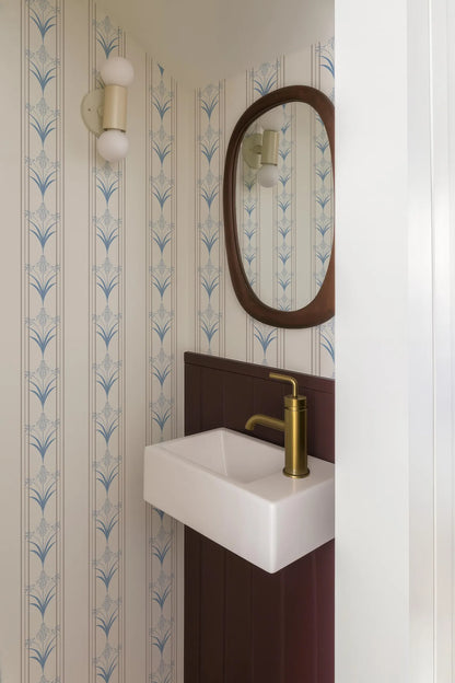 a bathroom with a white sink and a gold faucet, set against a wall with blue and white patterned wallpaper. There is a mirror above the sink, and a light fixture is mounted on the wall.