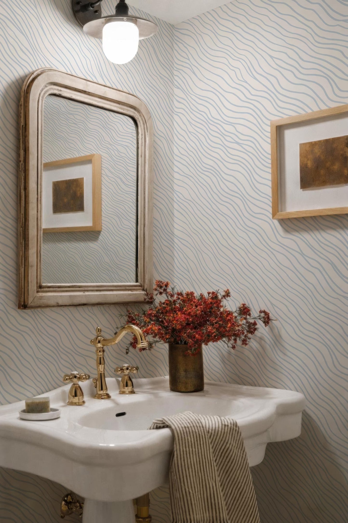 a bathroom with a white pedestal sink, a gold faucet, and a framed picture on the wall. The sink is positioned under a light fixture, and there is a vase of flowers on the counter. The walls have a wavy pattern, and there is a striped towel hanging on the side of the sink.