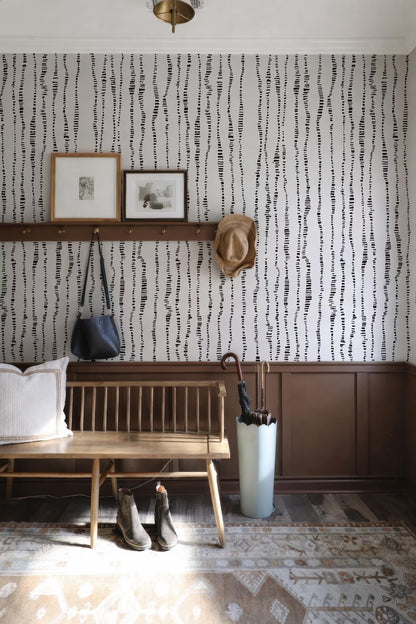a cozy entryway with a wooden bench, a hat rack, and a pair of boots. The wall behind the bench has a black and white striped pattern, and there are two framed pictures hanging on the wall. A white umbrella is placed on the floor next to the bench.