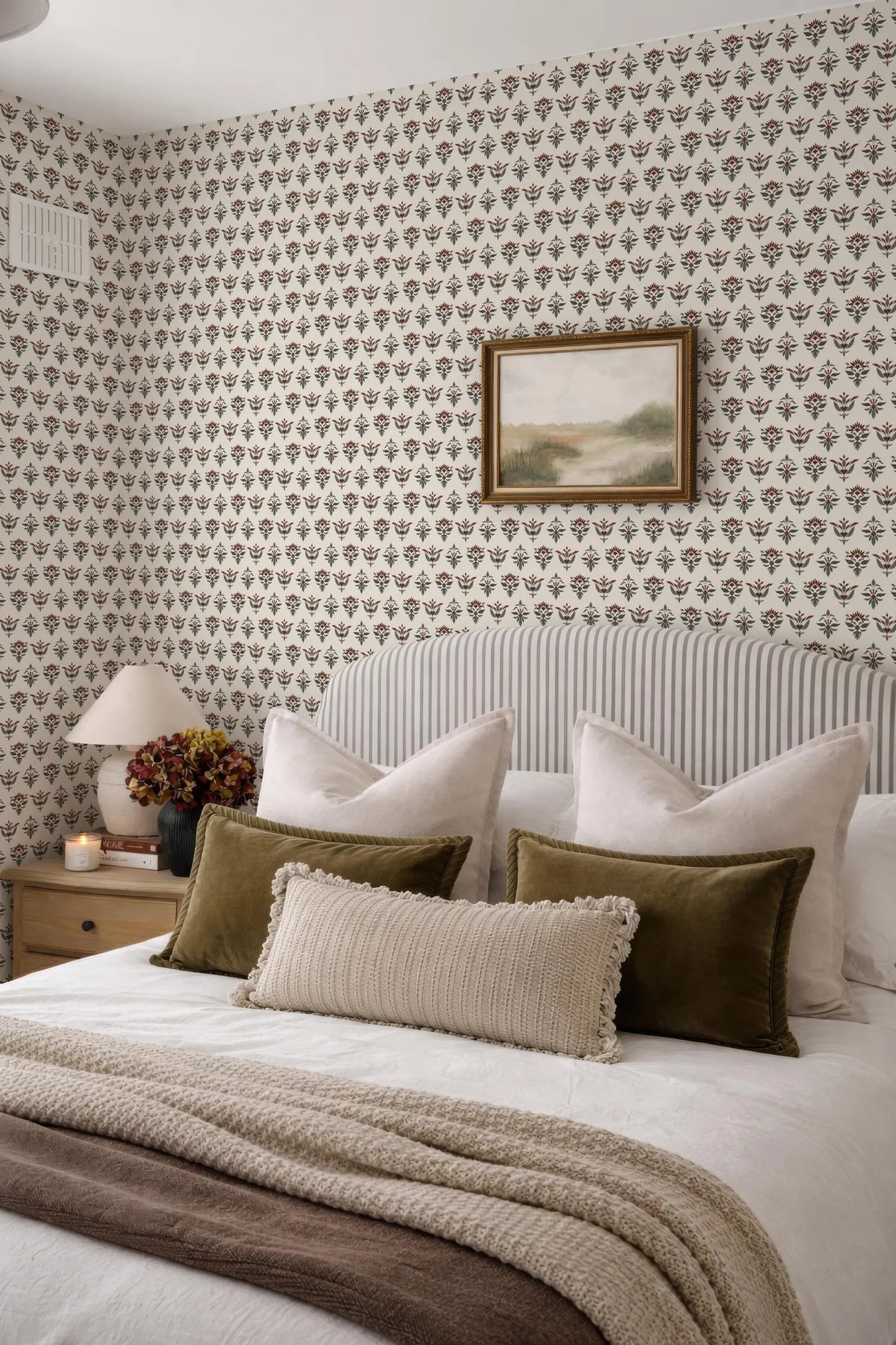 a cozy and inviting bedroom with a large bed adorned with a white comforter and a brown throw blanket. The bed is framed by a striped headboard and has several pillows in various shades of brown and beige. Above the bed, there is a framed landscape painting hanging on the wall. The room also features a nightstand with a lamp and a vase of flowers, adding to the overall aesthetic of the space.