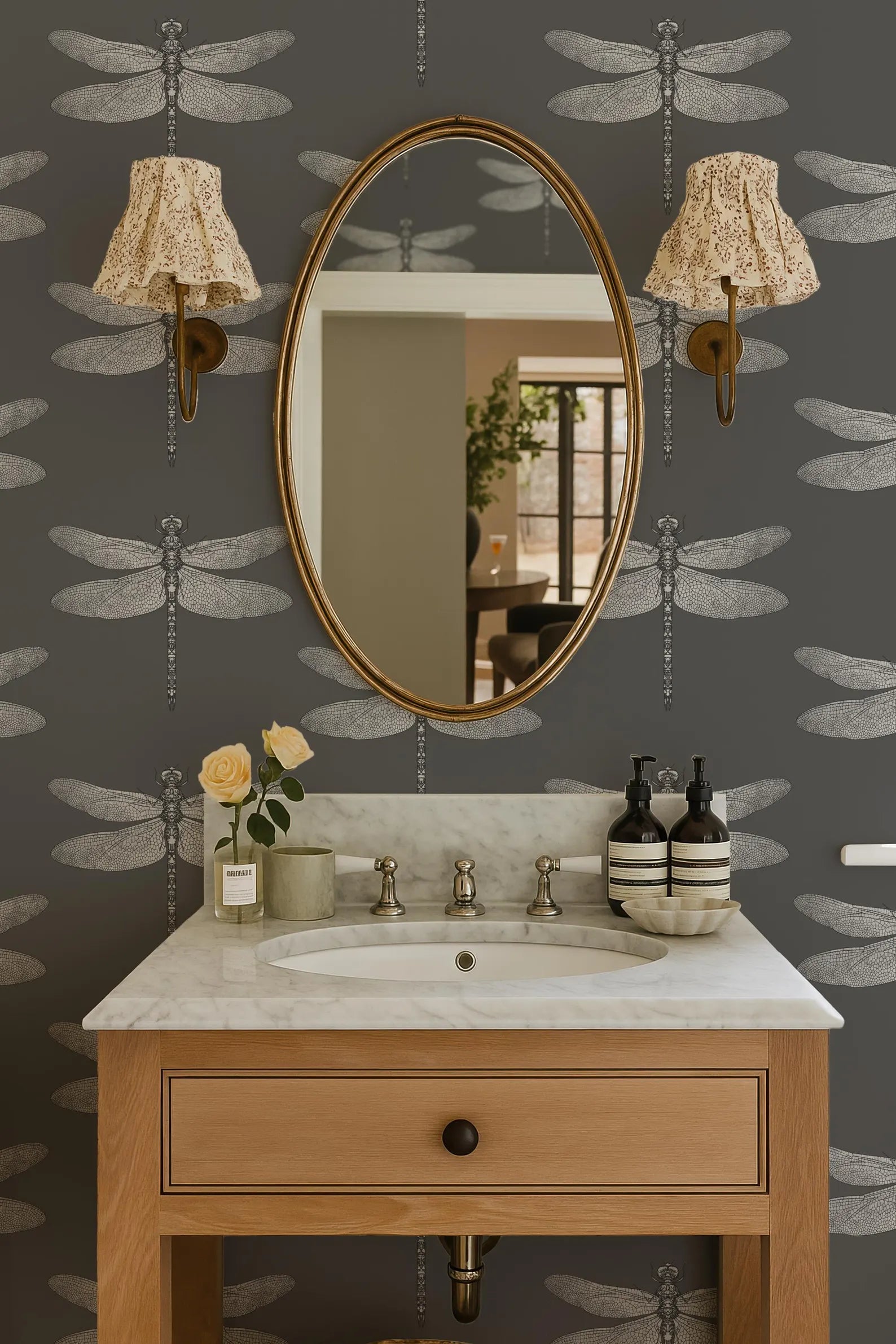a bathroom with a white marble sink, a wooden vanity, and a large oval mirror with gold frame. The walls are decorated with a dragonfly pattern wallpaper, and there are two lamps with white shades on either side of the mirror.