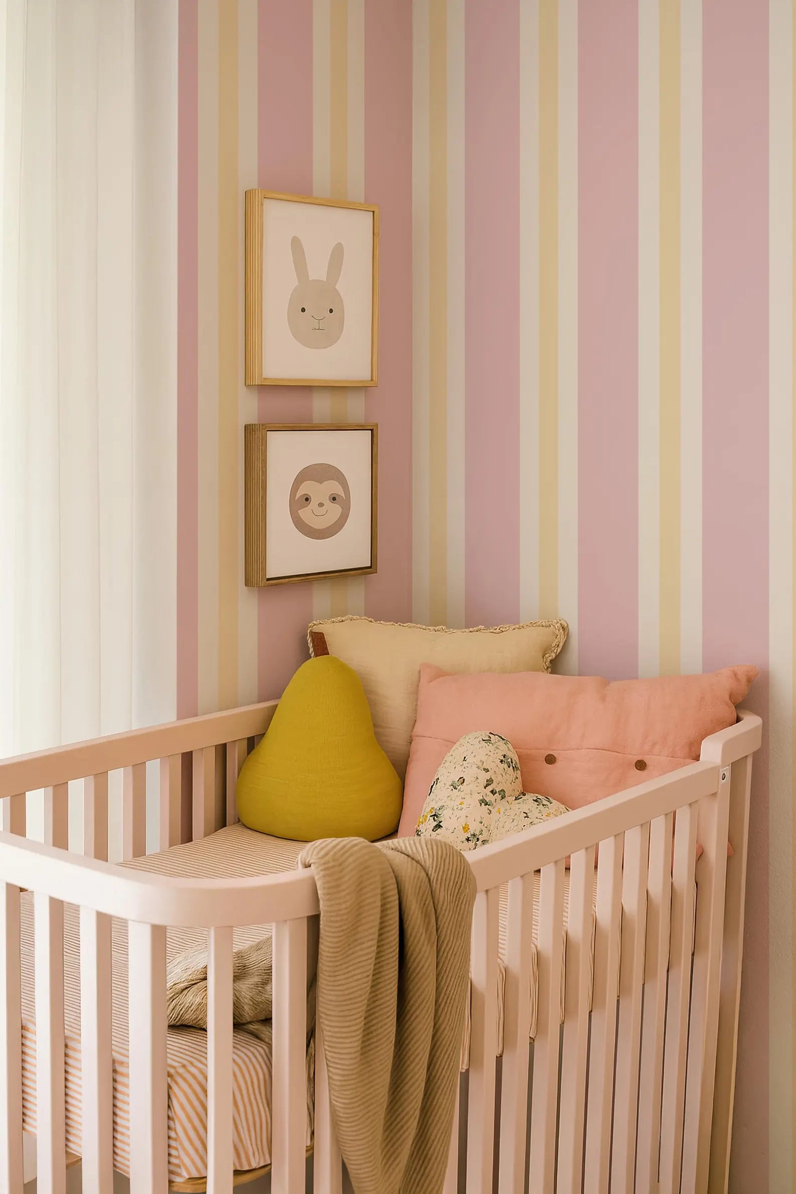 a cozy nursery with a white crib, adorned with a yellow pillow and a pink pillow. Above the crib, there are two framed pictures of animals, adding a playful touch to the room. The walls are decorated with pink and yellow striped wallpaper, creating a warm and inviting atmosphere.