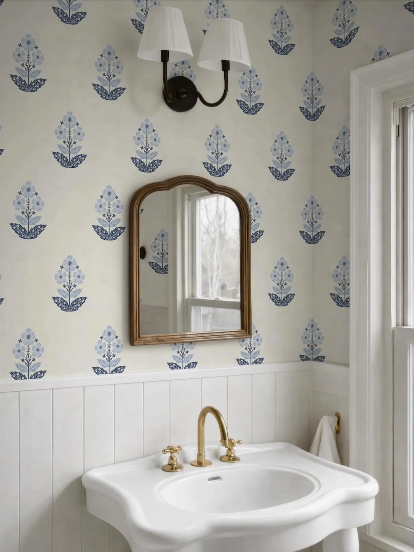 a bathroom with a white sink, a mirror, and a light fixture above it. The walls are covered in a patterned wallpaper featuring blue flowers.