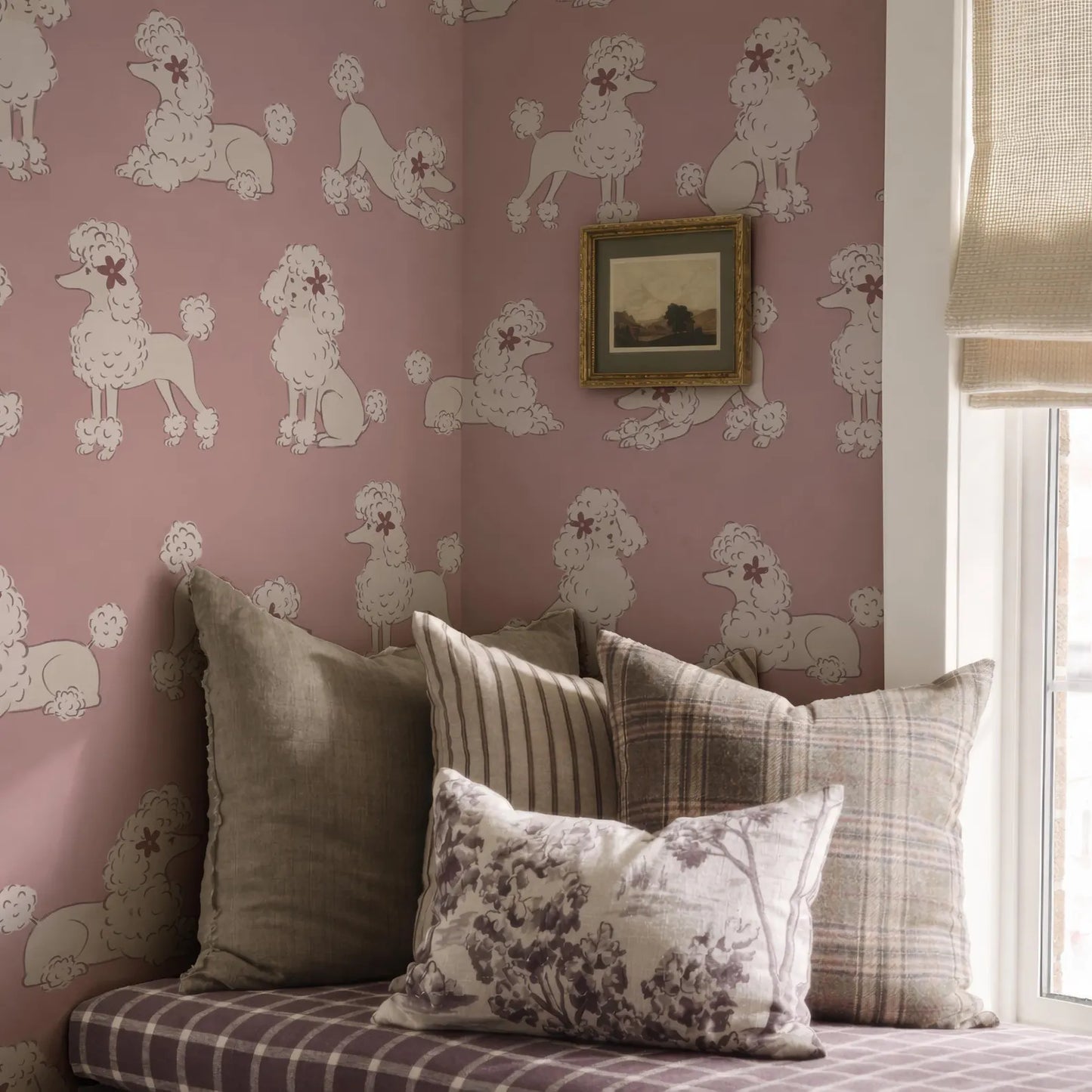 a pink wall with a repeating pattern of white poodle illustrations, creating a whimsical and playful atmosphere. In the foreground, there is a bed with a plaid comforter and several pillows, including one with a floral pattern. A framed picture is also visible on the wall, adding a personal touch to the room's decor.