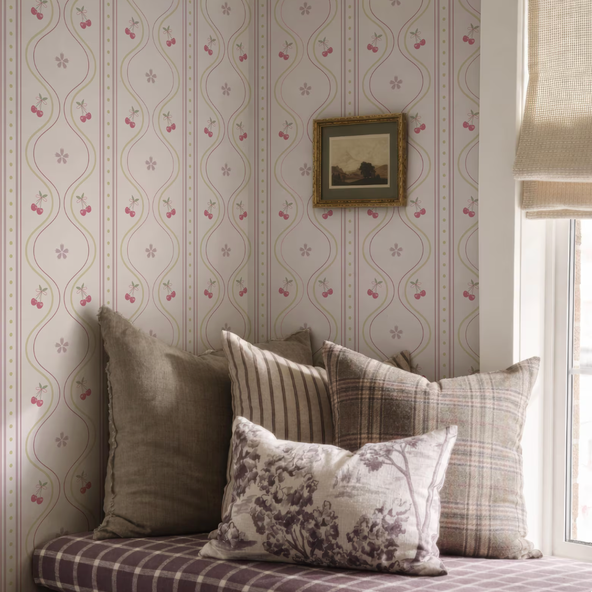 a cozy corner with a window, a wallpapered wall, and a framed picture. The wallpapered wall has a repeating pattern of pink and white flowers, and there are several pillows on a couch or bed in the foreground, creating a comfortable and inviting atmosphere.