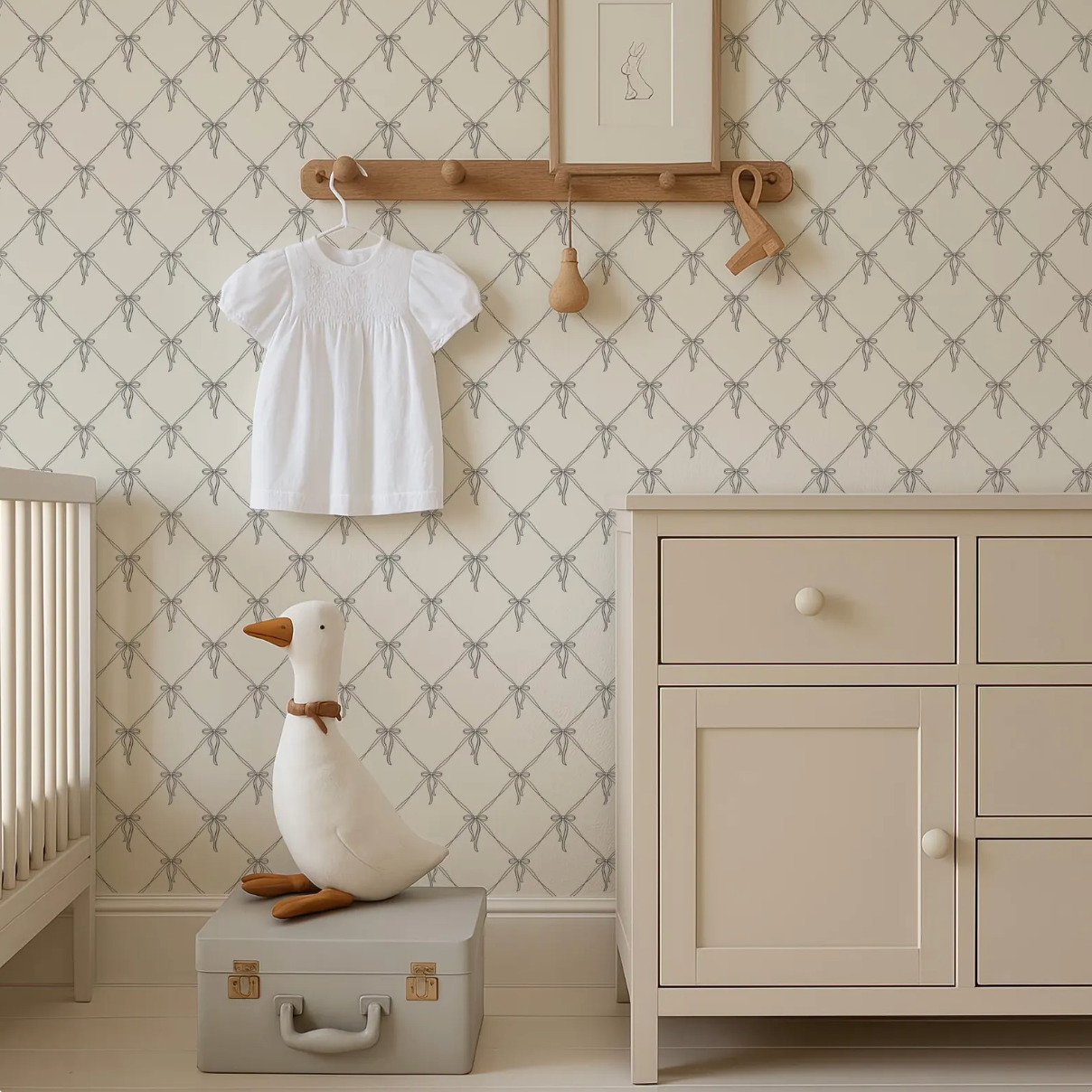 a nursery room with a white dresser, a wooden hanger, and a stuffed duck on a suitcase. The room has a patterned wallpaper and a wooden shelf with a picture frame.