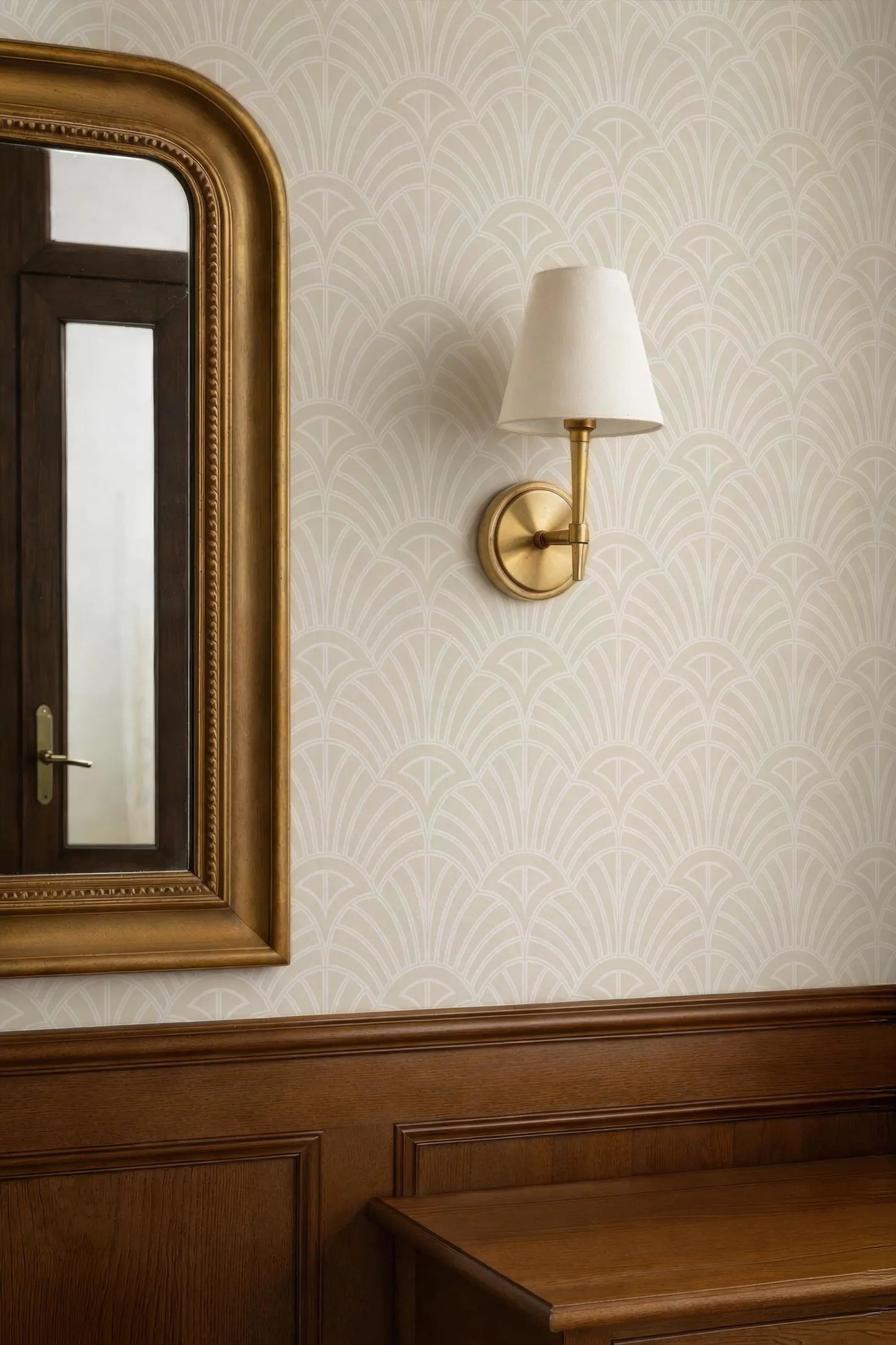 a wallpapered wall with a gold-framed mirror and a gold-colored wall lamp.
