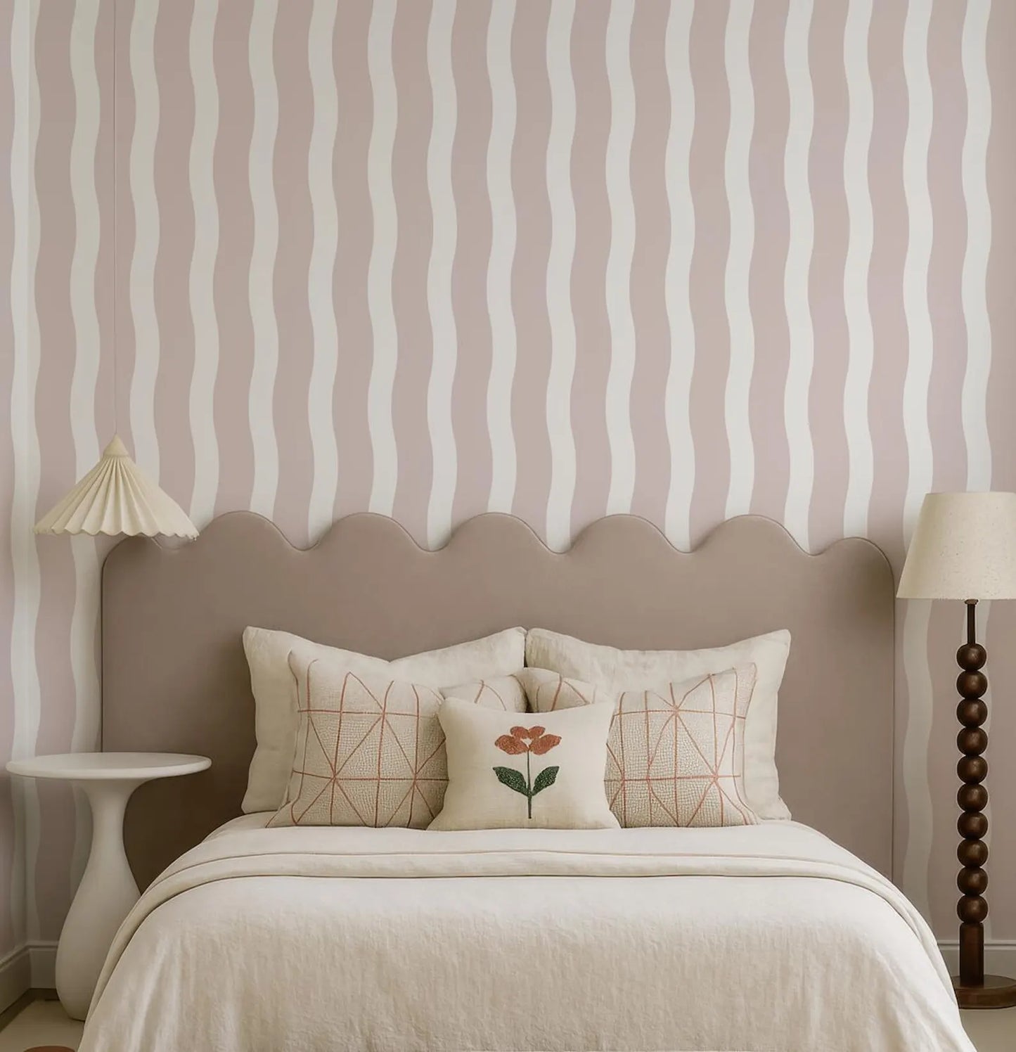 a well-decorated bedroom with a bed, a nightstand, and a lamp. The bed has a gray headboard and is adorned with a single red flower pillow. The walls are covered in a striped pattern, and the overall color scheme is neutral, creating a calm and inviting atmosphere.