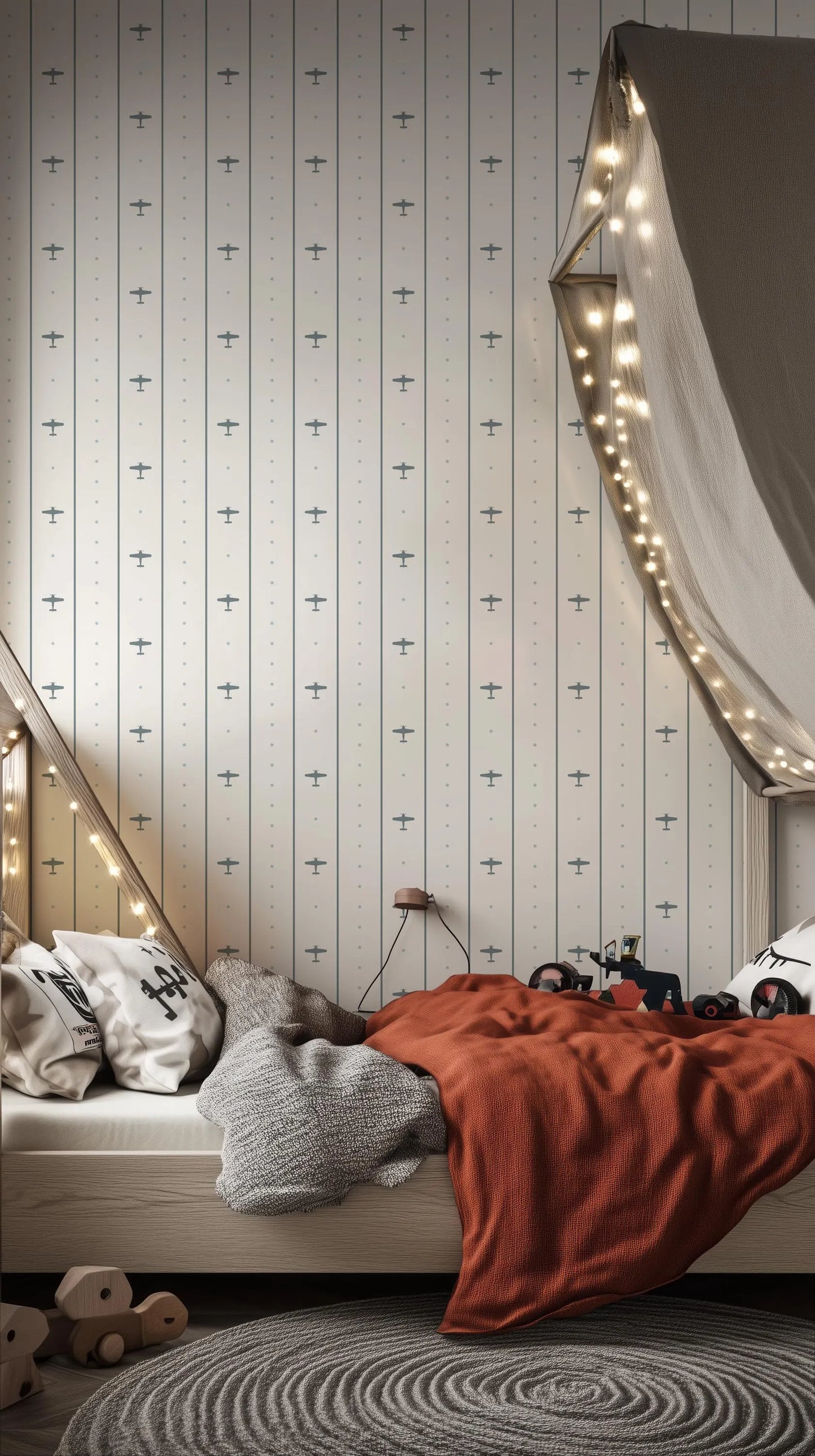a cozy bedroom scene with a bed, a gray blanket, and a red blanket. The bed is covered with a gray blanket and has a wooden headboard. The walls are decorated with a striped wallpaper featuring small airplane designs. The room also has a canopy and string lights, creating a warm and inviting atmosphere.