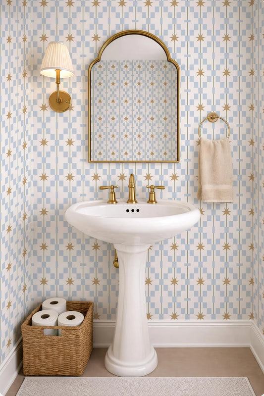 a bathroom with a white pedestal sink, a mirror, and a towel rack. The walls are covered in a patterned wallpaper with blue and gold stars. There is a basket of toilet paper on the floor next to the sink.
