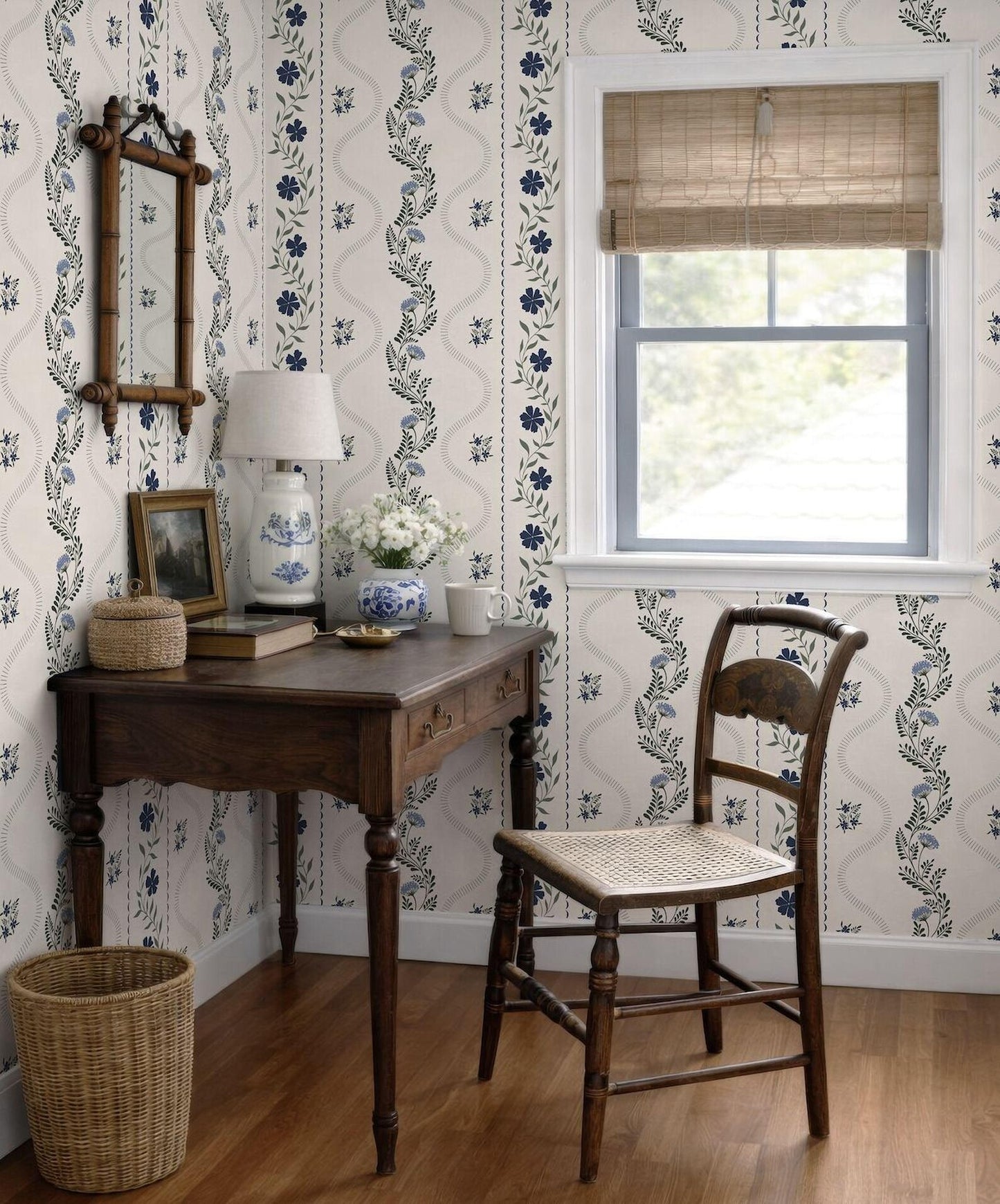 a cozy corner of a room with a wooden desk, a wicker chair, and a window with a bamboo blind. The room has a floral wallpaper pattern and a wooden floor.