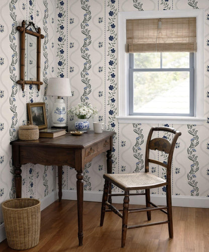 a cozy corner of a room with a wooden desk, a wicker chair, and a window with a bamboo blind. The room has a floral wallpaper pattern and a wooden floor.