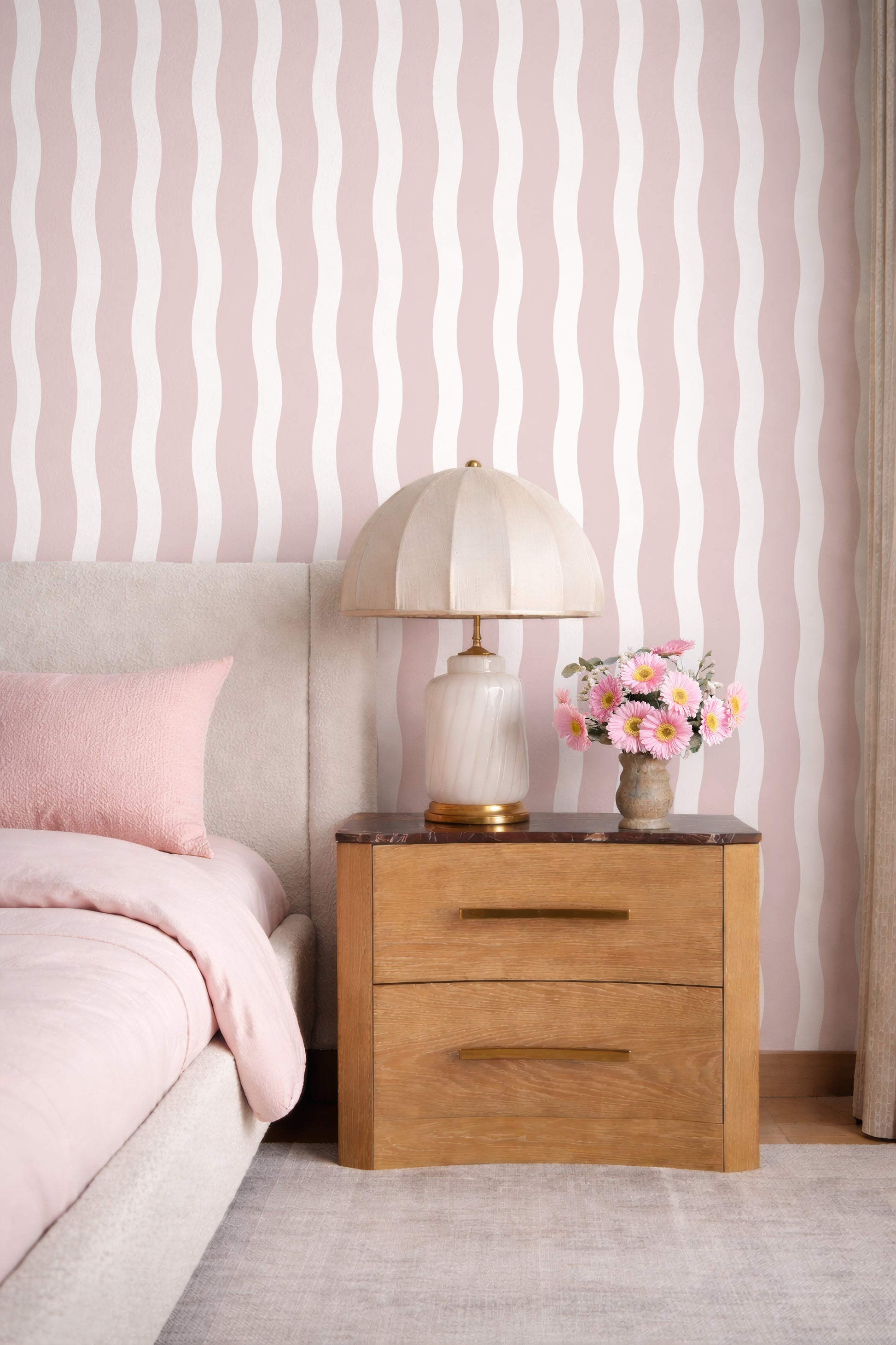 a bedroom with a bed, a wooden nightstand, and a lamp with a white shade. The wall behind the bed has a striped pattern in shades of pink and white. The nightstand has a marble top and two drawers. A vase with pink and white flowers is placed on the nightstand.