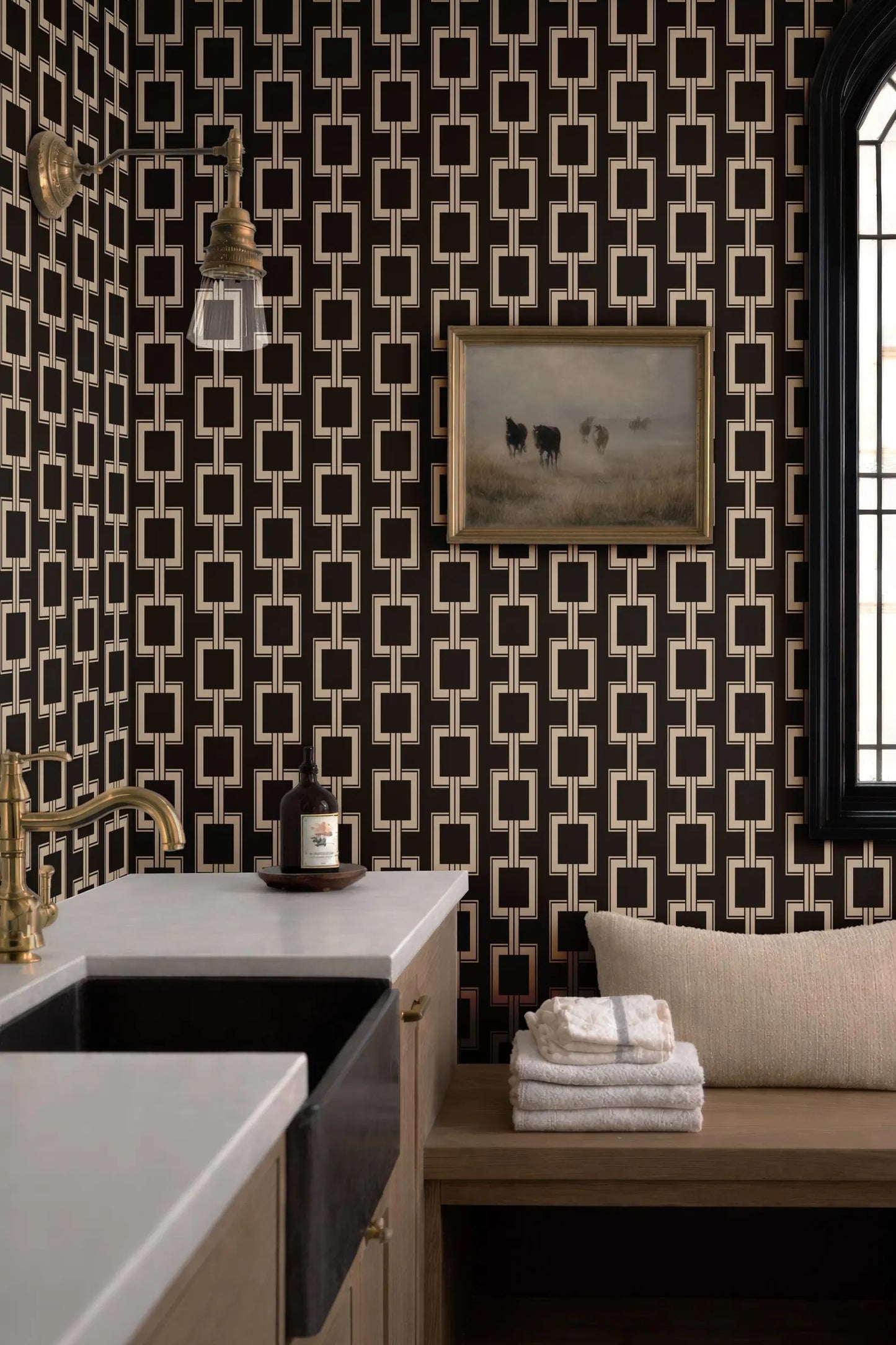 a modern bathroom with a black and white geometric wallpaper pattern, a white countertop, a sink, a framed painting of cows, and a wooden bench with a stack of towels on it.