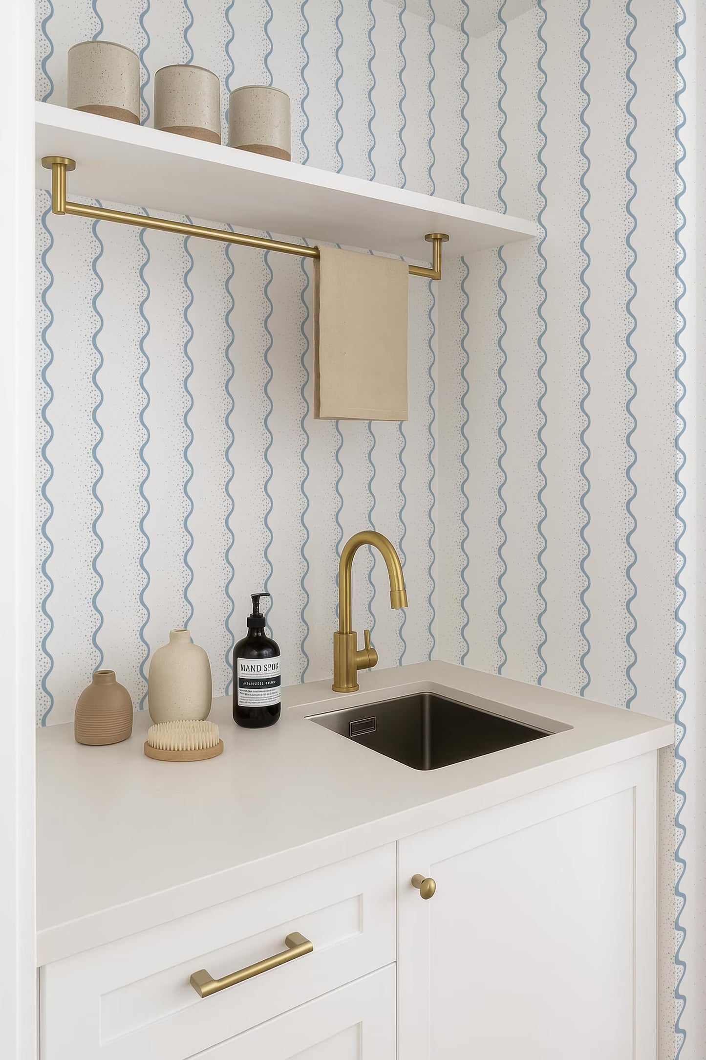 a bathroom with a white sink, a shelf above it holding three beige cups, and a gold faucet. The walls are covered in a blue and white striped wallpaper, and there is a white cabinet below the sink.