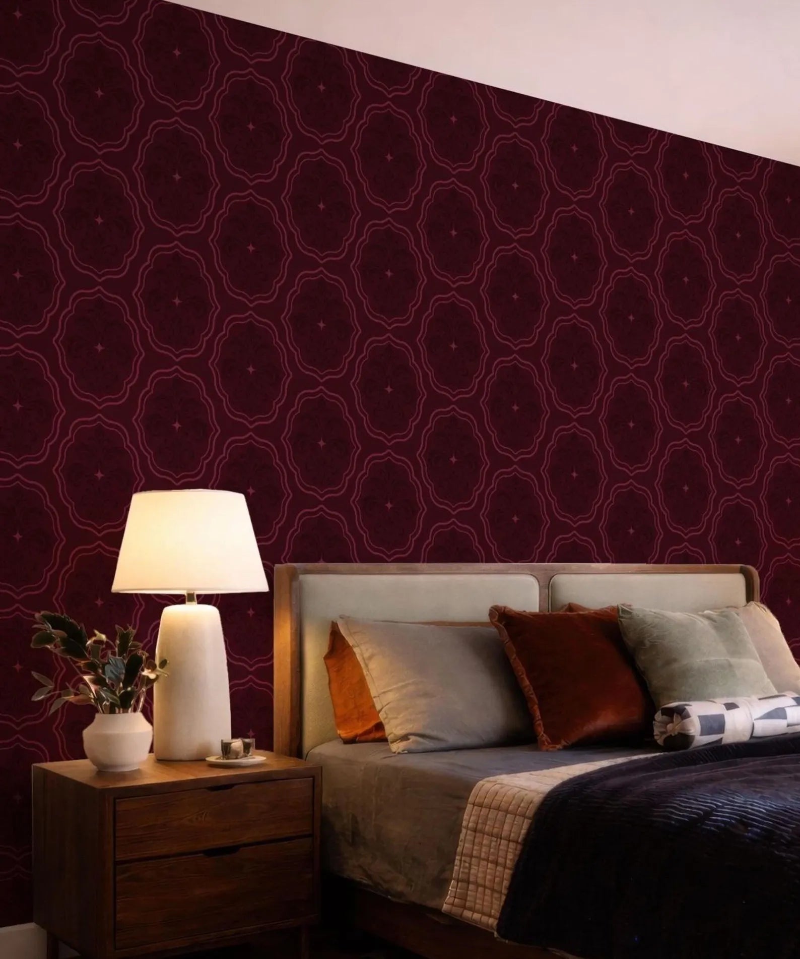 a bedroom with a bed, a nightstand, and a lamp. The walls are covered in a patterned wallpaper in a burgundy color.