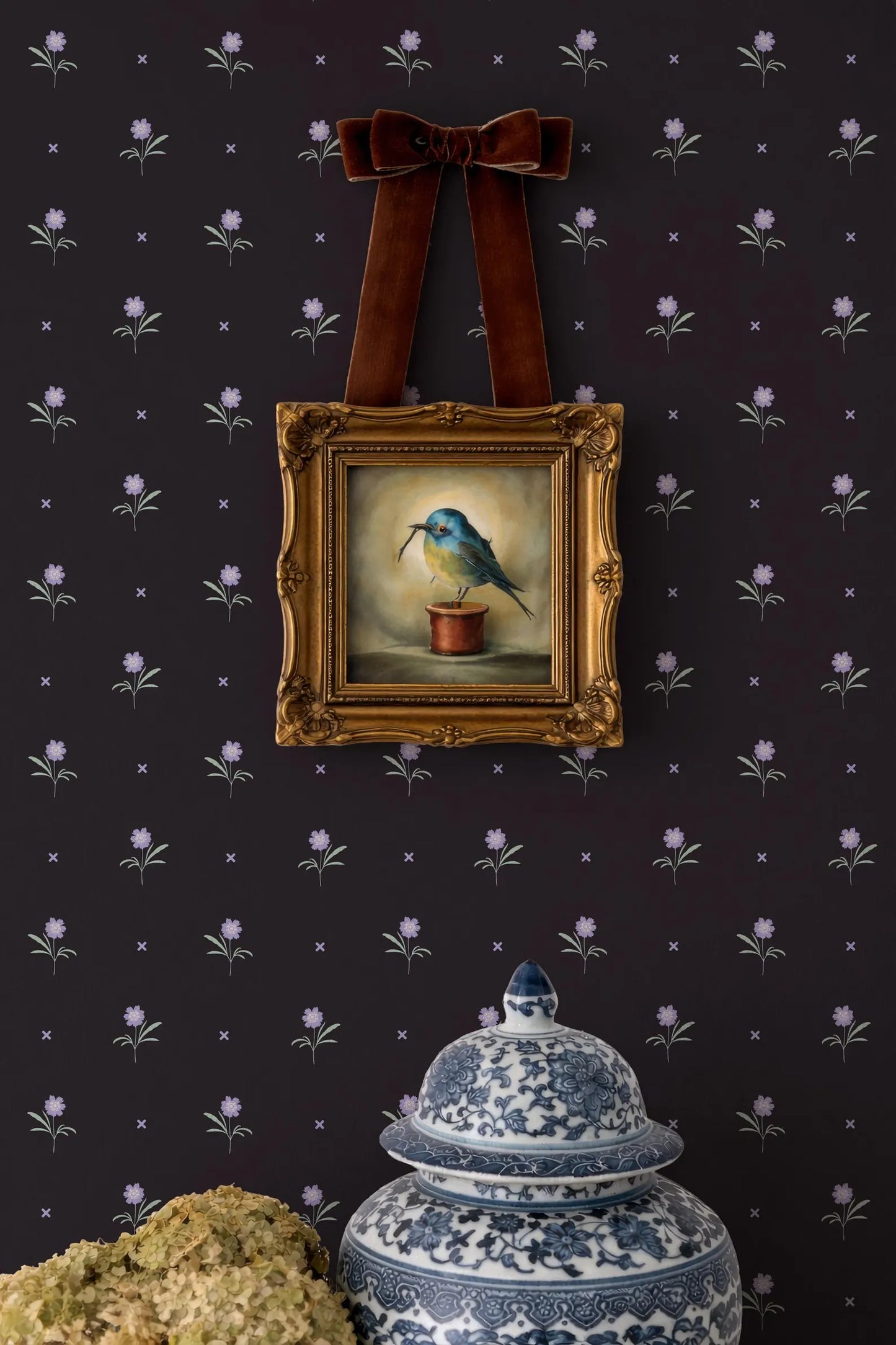 a wall with a dark purple floral wallpaper, a gold-framed painting of a blue bird on a pot, and a blue and white porcelain urn with a lid.