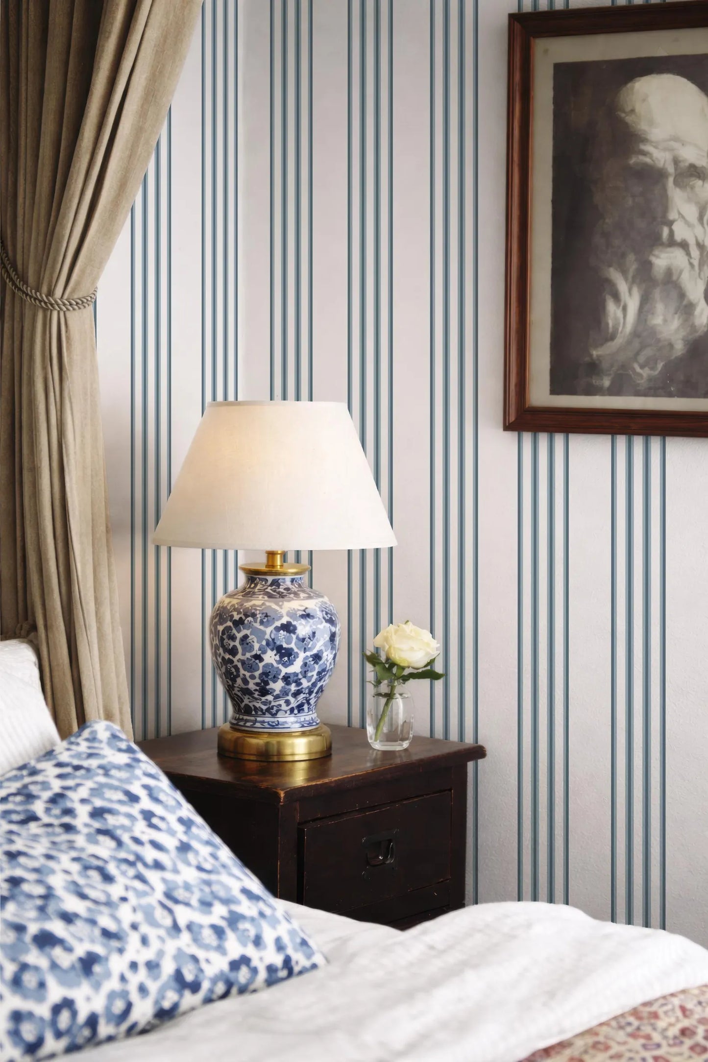 a bedroom with a bed, a nightstand, and a lamp with a white shade. The wall behind the nightstand has a striped wallpaper pattern. A framed picture is hanging on the wall, and a vase with a single white flower is placed on the nightstand.
