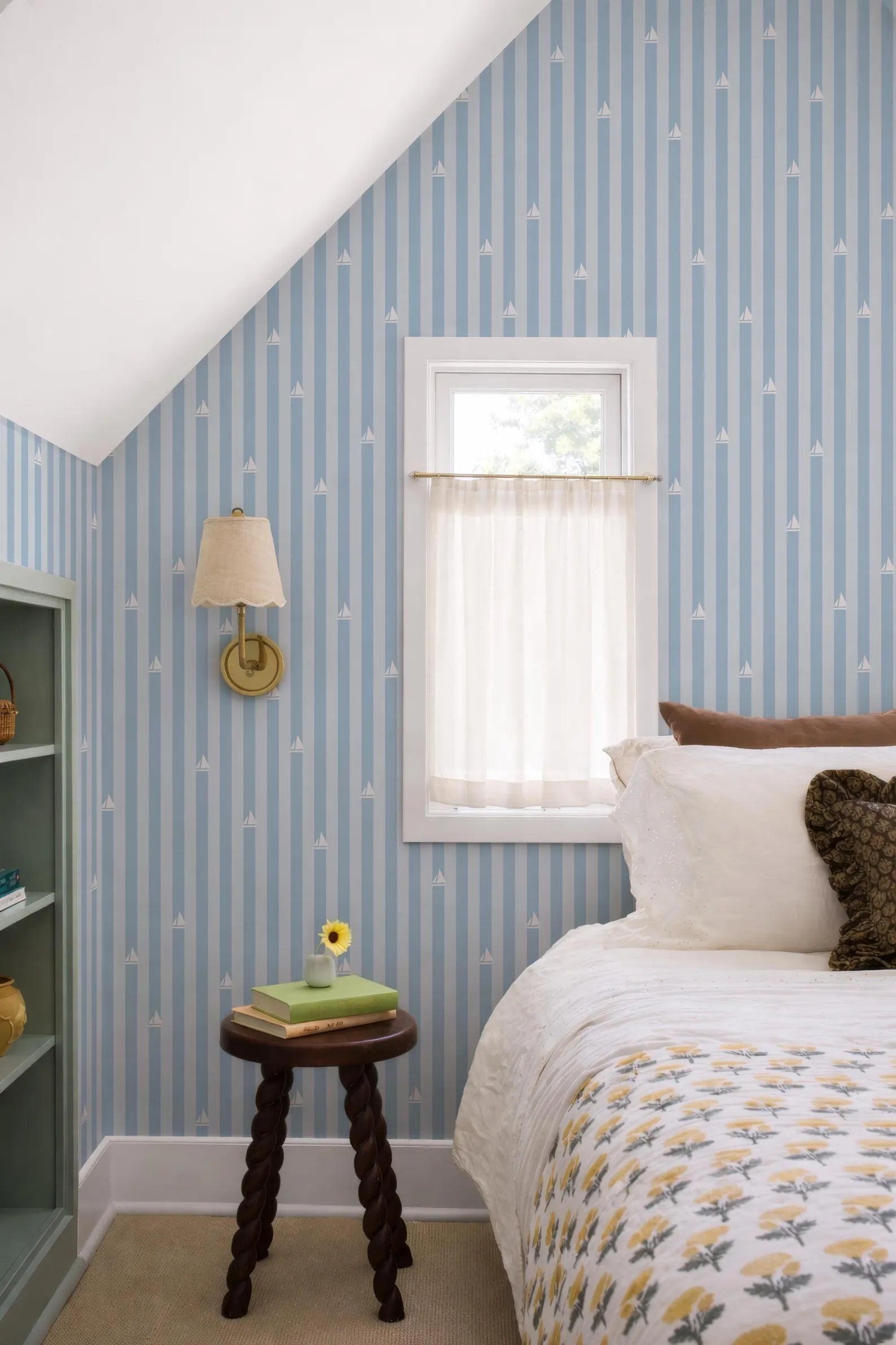 a cozy bedroom with a bed, a small wooden table, and a window with a white curtain. The walls are covered in blue and white striped wallpaper, and there is a small table with a lamp and a vase of flowers.