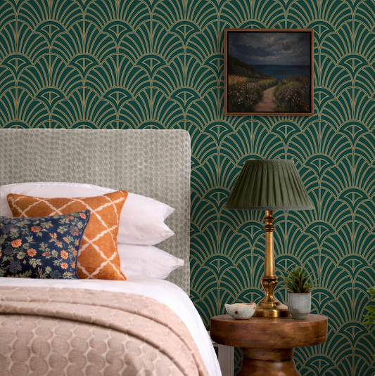 a bedroom with a bed, a lamp, and a framed landscape painting on the wall. The bed has a white headboard and is adorned with colorful pillows. The lamp is placed on a wooden side table, and the wall behind the bed is covered in a patterned wallpaper.