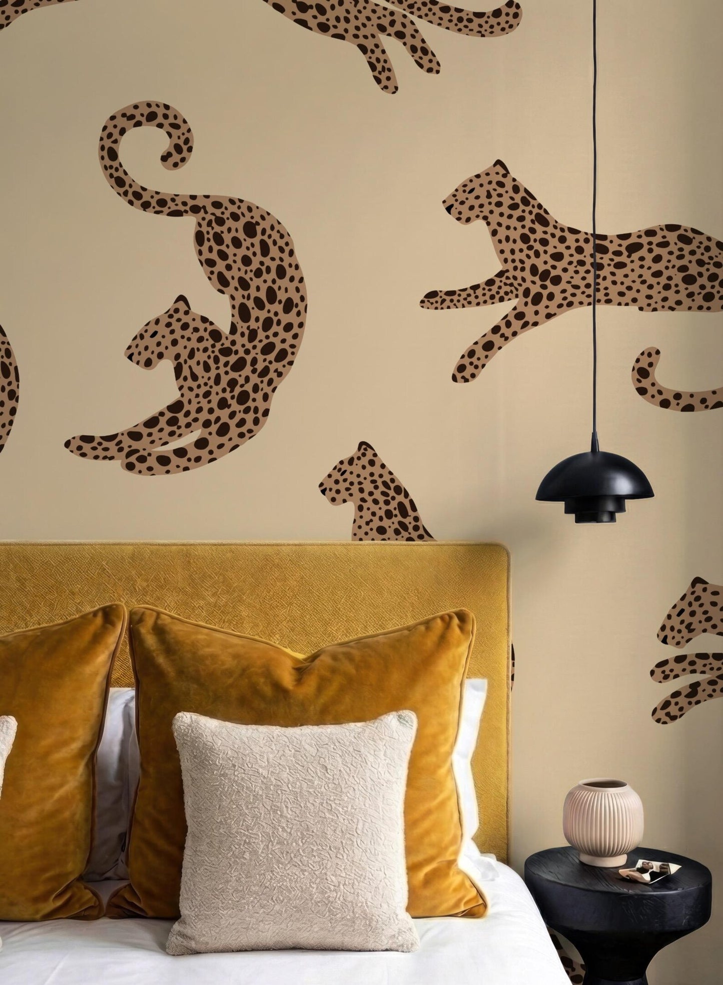 a bedroom with a yellow headboard and a wallpapered wall with a repeating pattern of leopard silhouettes. The room has a cozy and stylish atmosphere, with a bed adorned with yellow and white pillows, a nightstand, and a hanging light fixture.