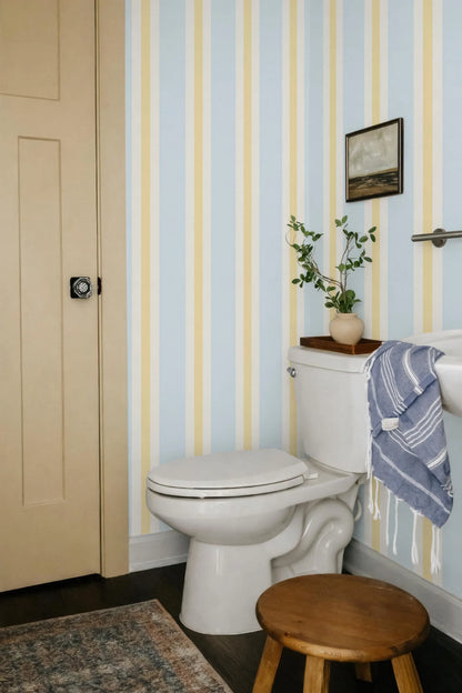 a bathroom with a white toilet, a wooden stool, and a blue and white striped towel hanging on a rack. The walls are covered in blue and yellow striped wallpaper, and there is a small plant on the toilet tank.