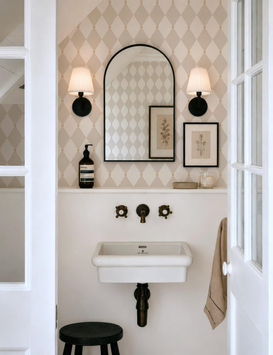 a small bathroom with a white sink, a mirror, and two wall sconces. The walls are covered in a diamond patterned wallpaper, and there is a stool in the corner.