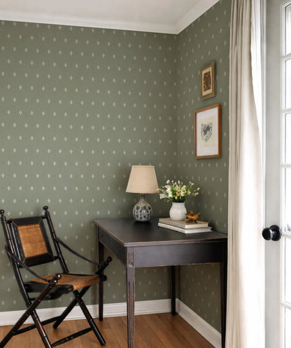 a cozy corner of a room with a wooden desk, a lamp, and a vase of flowers. The room has a green and white patterned wallpaper, and there is a wooden chair in the corner.
