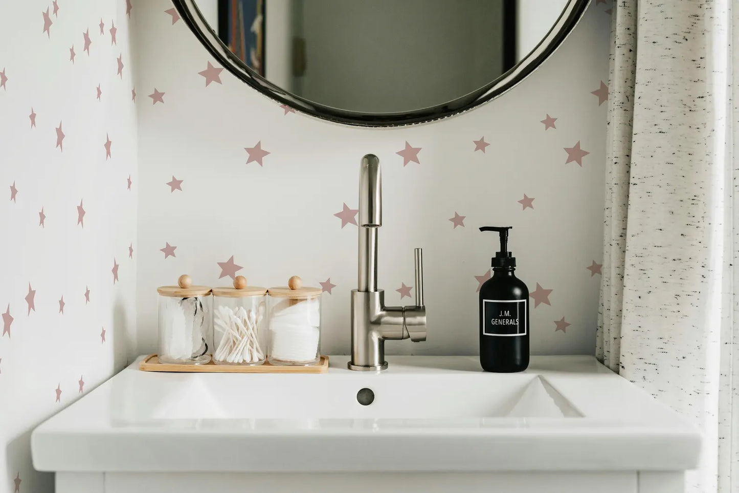 a bathroom sink with a silver faucet, a black soap dispenser, and a wooden tray with cotton swabs and cotton buds. The sink is set against a wall with pink stars on it, and there is a round mirror above the sink.