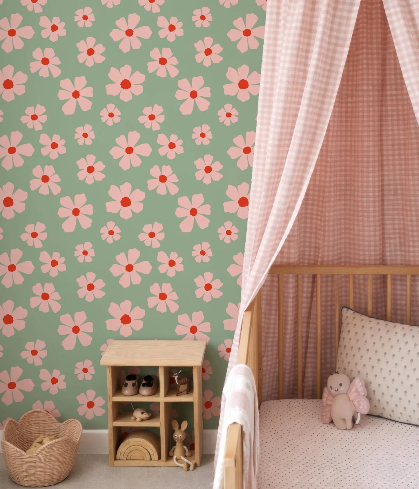 a nursery room with a green wall adorned with pink and white flower patterns. A wooden crib is situated next to the wall, and a basket is placed on the floor. A small wooden shelf is also present in the room, housing various toys.