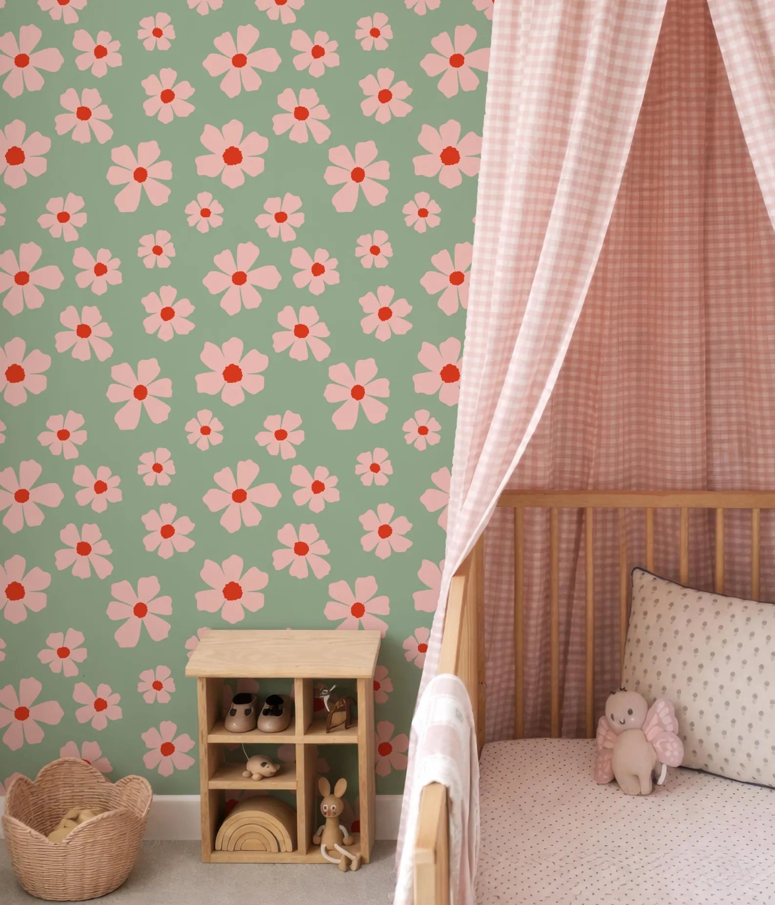 a nursery room with a green wall adorned with pink and white flower patterns. A wooden crib is situated next to the wall, and a basket is placed on the floor. A small wooden shelf is also present in the room, housing various toys.