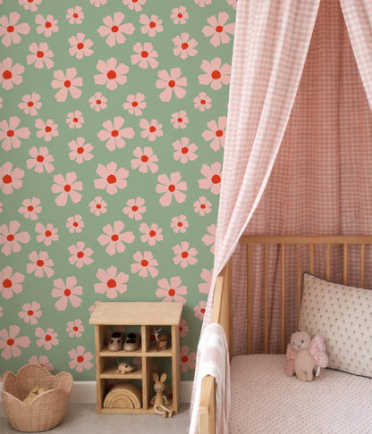 a nursery room with a green wall adorned with pink and white flower patterns. A wooden crib is situated next to the wall, and a basket is placed on the floor. A small wooden shelf is also present in the room, housing various toys.