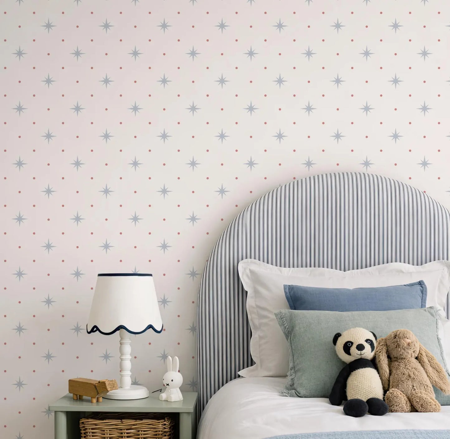 a cozy bedroom scene with a bed, a nightstand, and a stuffed animal. The bed has a striped headboard and is adorned with blue and white pillows. A stuffed panda bear and a stuffed rabbit are resting on the bed, adding a playful touch to the room. The nightstand has a lamp and a small toy car, providing a functional and decorative element to the space. The wall behind the bed is decorated with a pattern of stars and dots, creating a starry atmosphere in the room.
