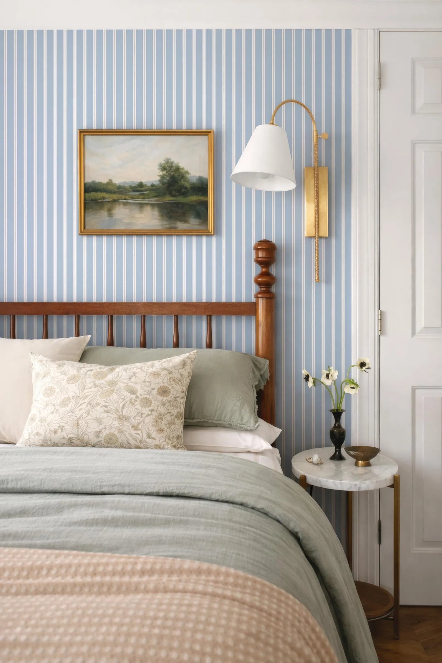 a cozy bedroom with a wooden headboard, a blue and white striped wallpaper, and a painting on the wall. The bed is adorned with a gray comforter and a white pillow, and a small side table with a vase of flowers is situated next to the bed.