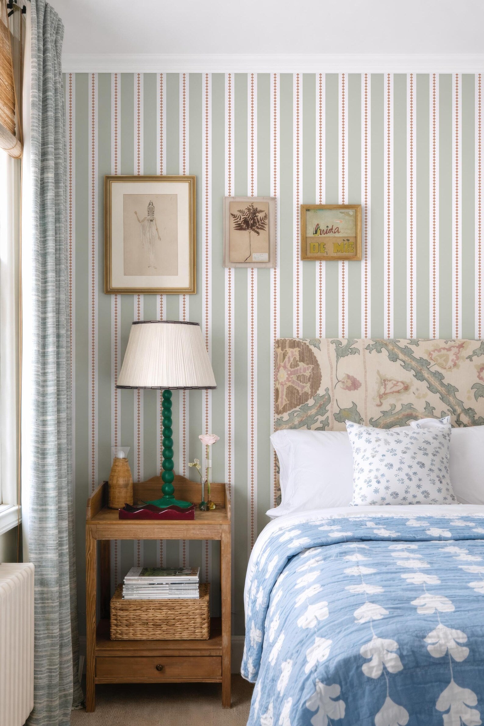 a cozy bedroom with a bed, a nightstand, and framed artwork on the wall. The bed has a blue and white patterned comforter and a floral headboard. The nightstand has a lamp and a basket of books. The wall is decorated with framed artwork, including a painting and a photograph.