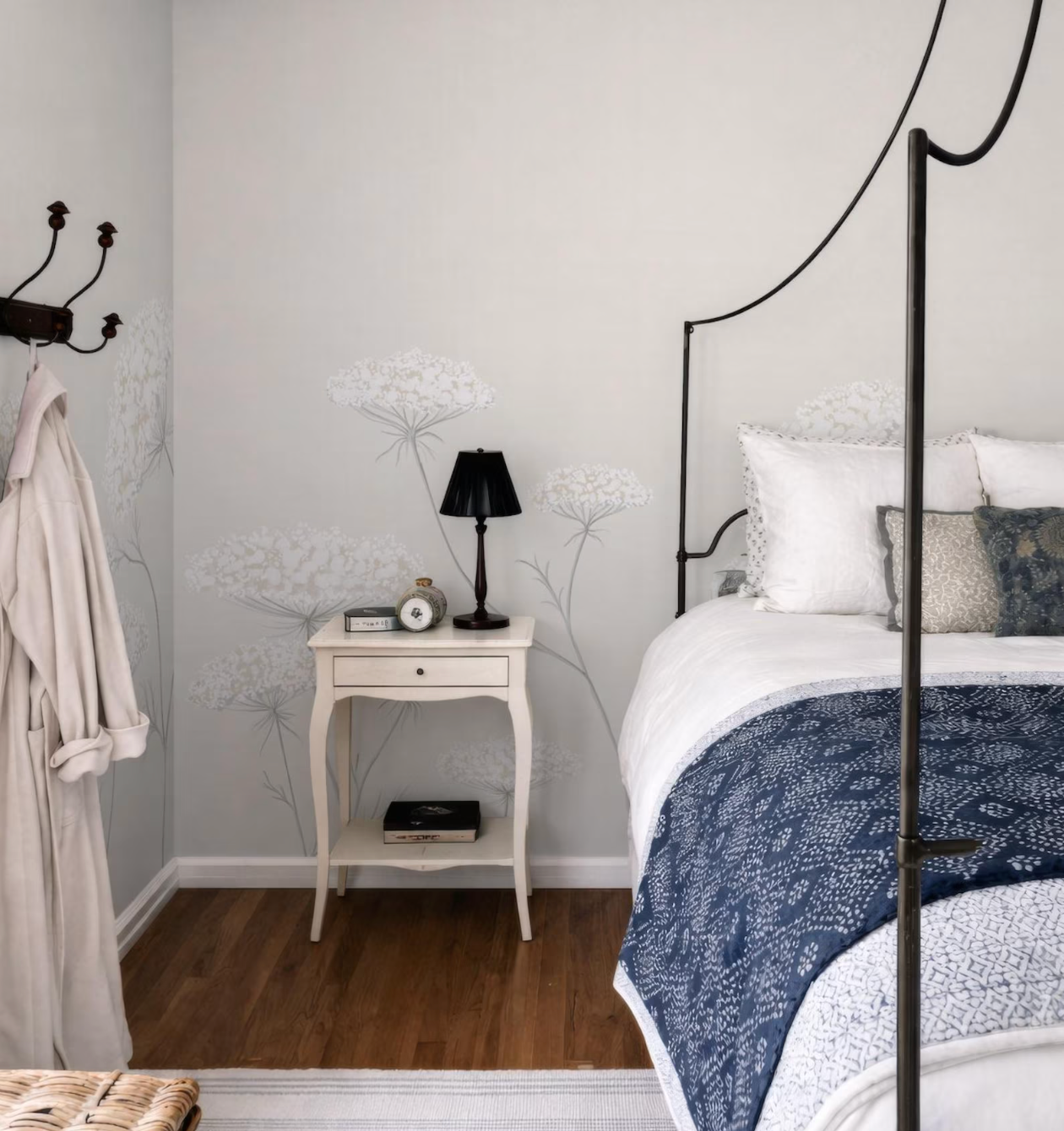 a cozy bedroom with a white bed frame, a nightstand, and a lamp. The room has a neutral color scheme with white walls and a blue and white patterned bedspread.