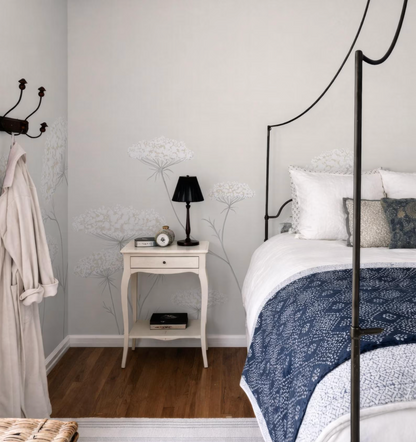 a cozy bedroom with a white bed frame, a nightstand, and a lamp. The room has a neutral color scheme with white walls and a blue and white patterned bedspread.