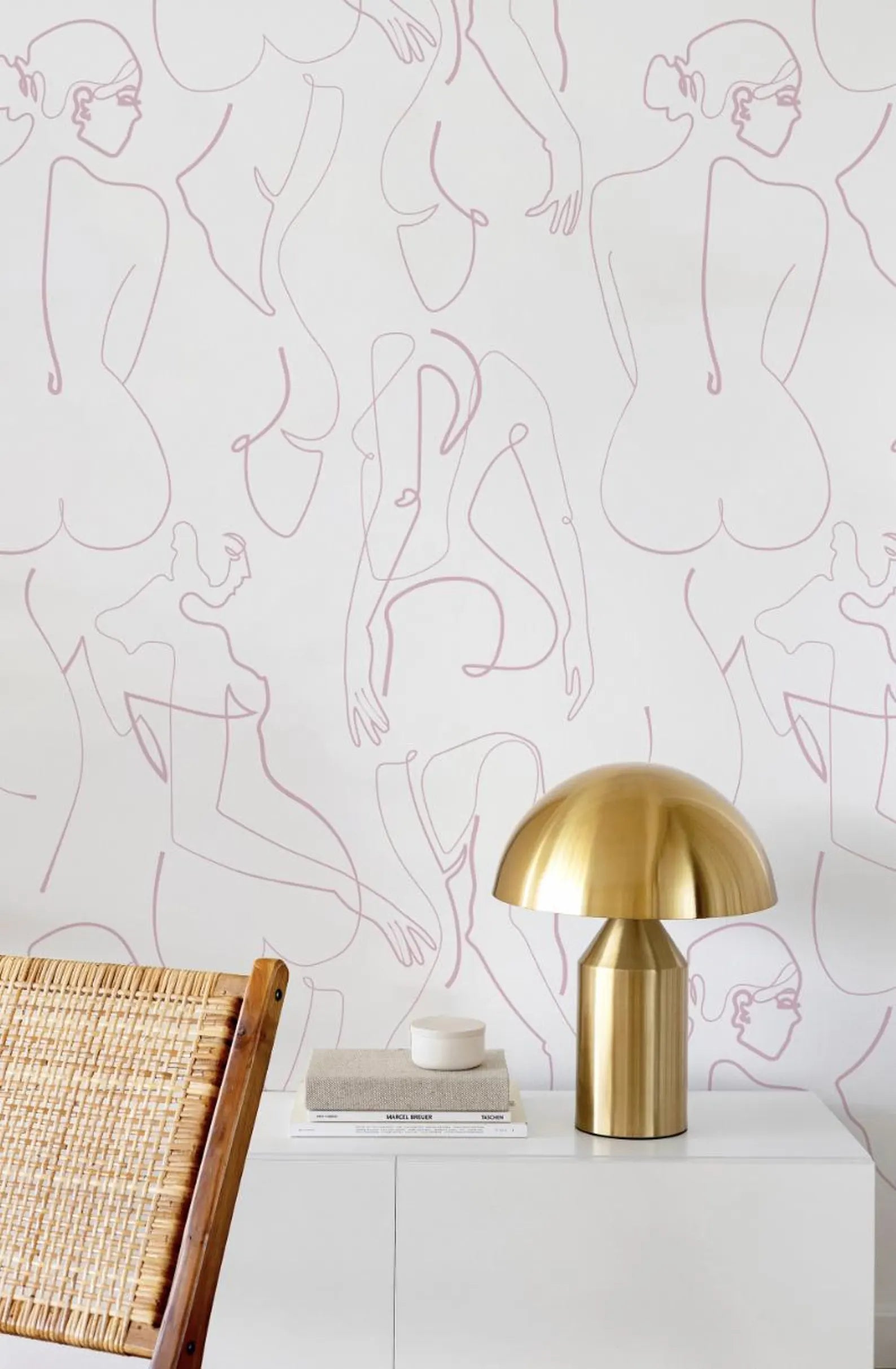 a white table with a brass lamp on top, a woven chair, and a stack of books. The background is a wall with a repeating pattern of abstract human figures in pink and white.