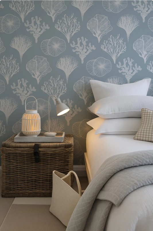 a bedroom with a bed, a wicker side table, and a lamp. The wall behind the bed has a pattern of white and blue floral designs.