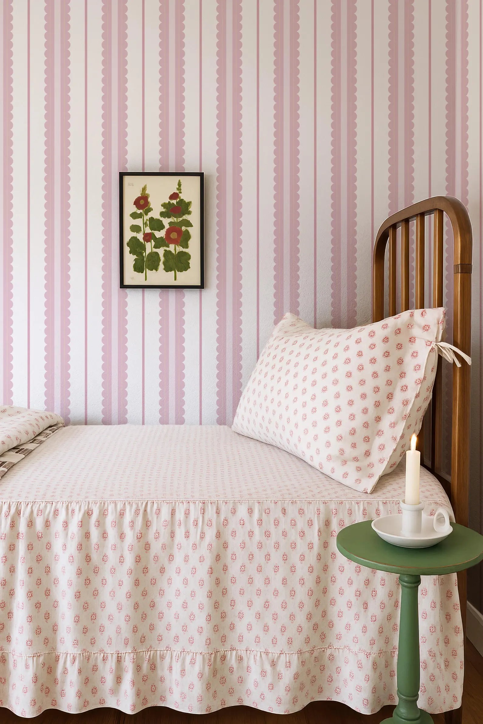 a cozy bedroom with a bed, a wooden headboard, and a framed floral print on the wall. The bed is covered in a white and pink patterned bedspread, and there is a green side table next to the bed with a white candle on it.