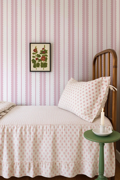 a cozy bedroom with a bed, a wooden headboard, and a framed floral print on the wall. The bed is covered in a white and pink patterned bedspread, and there is a green side table next to the bed with a white candle on it.