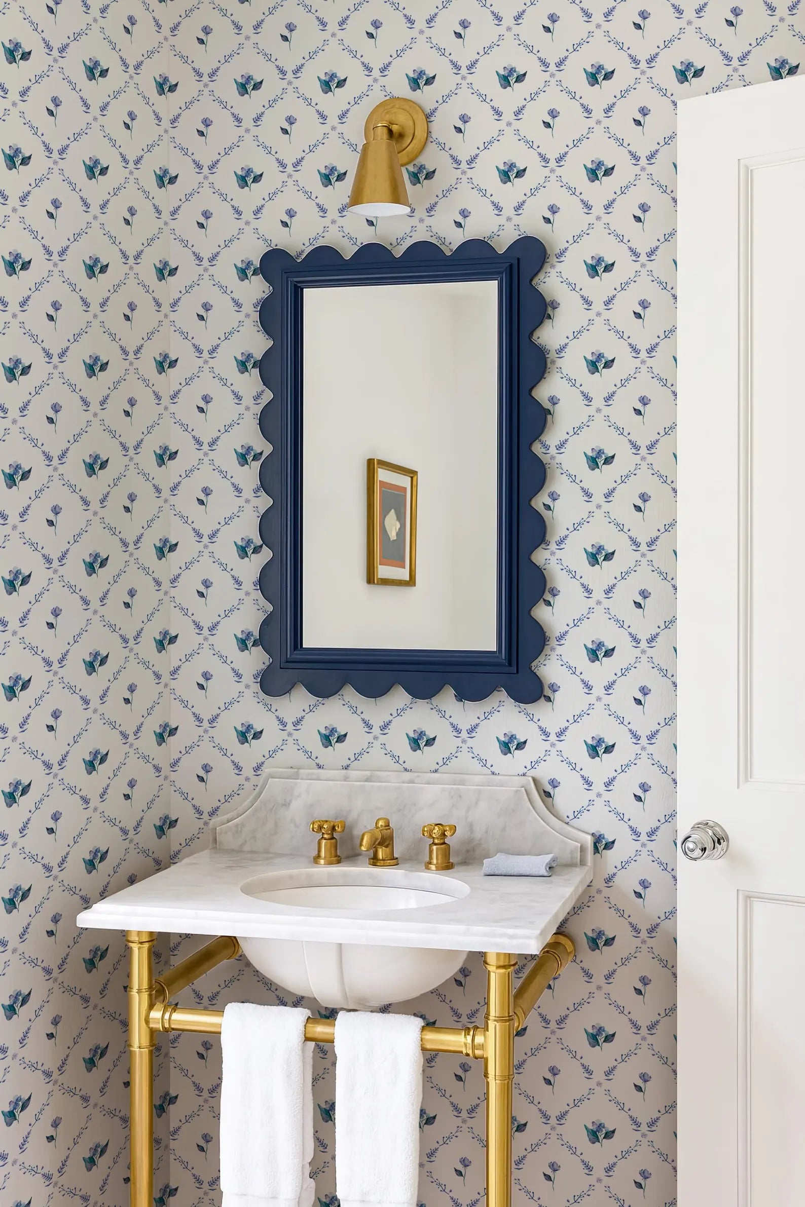 a bathroom with a white sink, a blue scalloped mirror, and a gold faucet. The walls are covered in blue and white floral wallpaper, and there is a white door in the background.