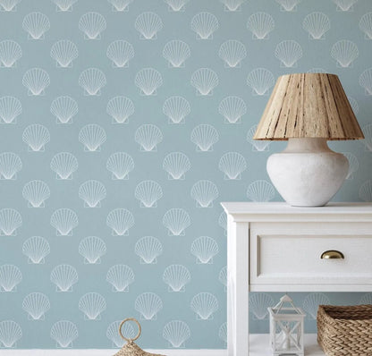 a light blue wall with a repeating pattern of white seashells, creating a coastal or nautical theme. A white table with a lamp and a woven basket are placed against the wall, adding a touch of home decor to the scene.