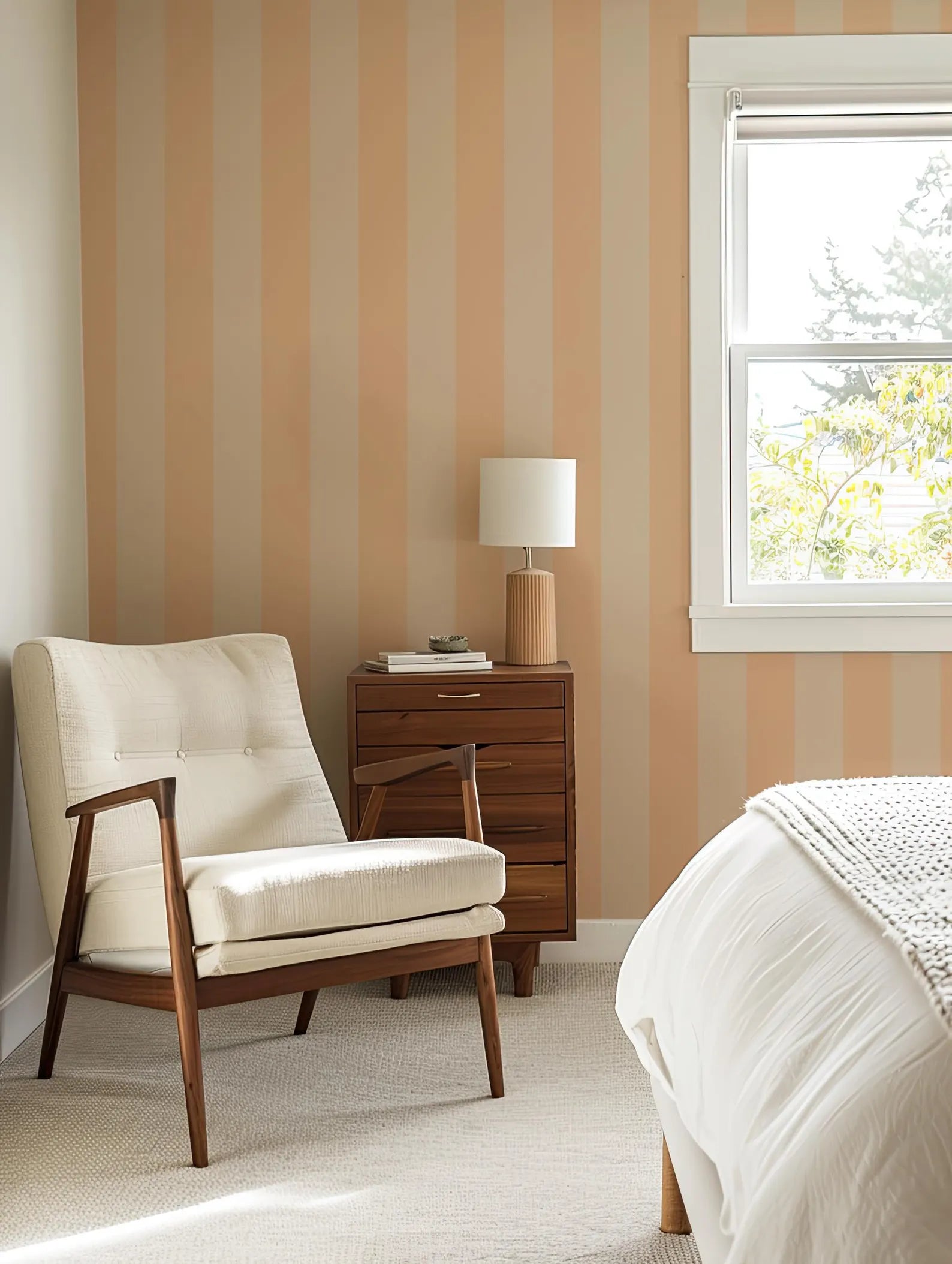 a cozy bedroom with a white armchair, a wooden side table with a lamp, and a window overlooking a tree outside.