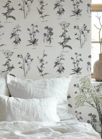 a bedroom setting with a white bed and a wallpapered wall with black and white botanical illustrations.