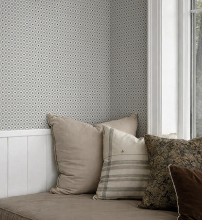 a cozy corner of a room with a white wall featuring a patterned wallpaper. The corner is decorated with a variety of pillows, including a beige pillow, a striped pillow, and a brown floral pillow, arranged on a couch or bed.