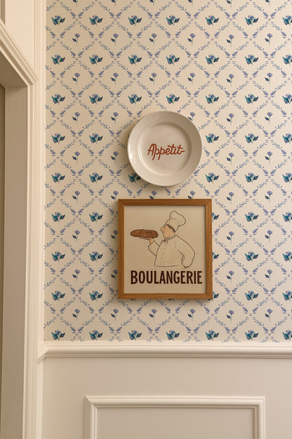 a wall with a framed picture of a chef holding a loaf of bread, a plate with the word "Appétit" written on it, and a wallpapered wall with a floral pattern.